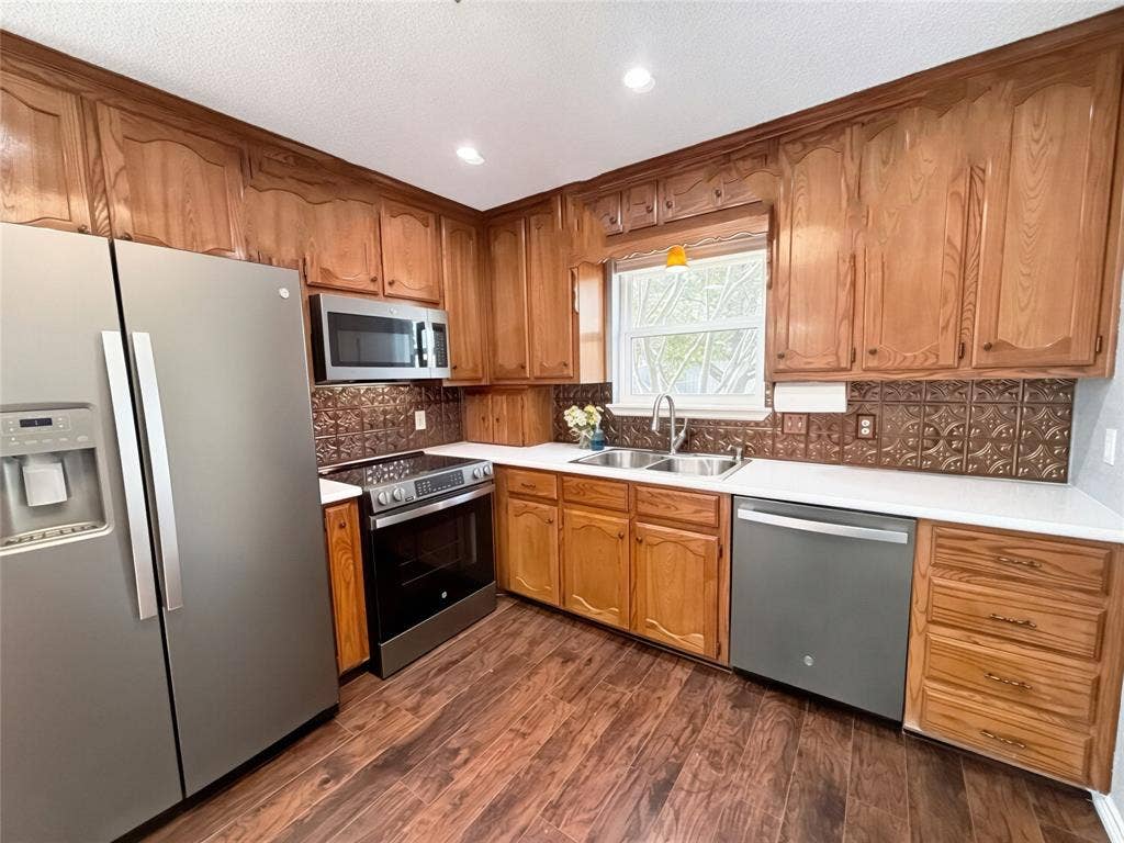 Kitchen featuring stained wood cabinetry, stainless steel appliances, patterned metal tile backsplash, white countertops, and wood-finish flooring