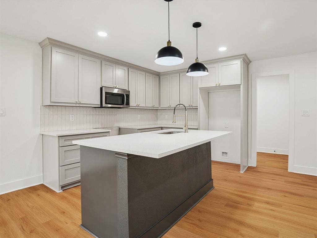 9059 Quinn Street, Dallas, TX 75217 Kitchen featuring light gray cabinetry, solid-surface countertops, a center island with a built-in sink, wood-finish flooring, and two dome-style pendant lights