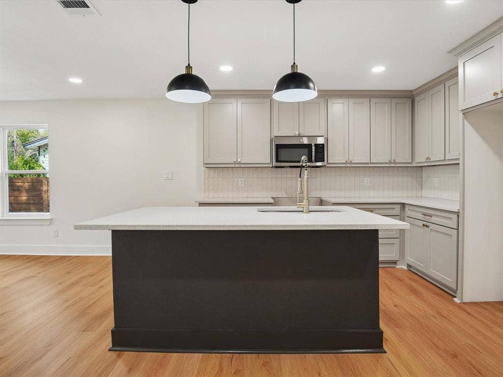 9059 Quinn Street, Dallas, TX 75217 Kitchen featuring a central island with a solid surface countertop, light-toned cabinetry, stainless steel microwave, vertical-tile backsplash, and dual pendant lighting over wood-finish flooring