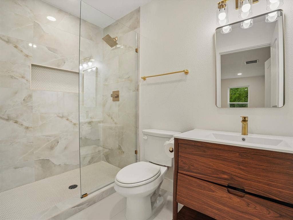 9059 Quinn Street, Dallas, TX 75217 Bathroom featuring a wood-finish vanity with a white countertop, walk-in shower with large-format tile walls and a built-in recessed niche, gold-toned hardware, and a modern rectangular mirror