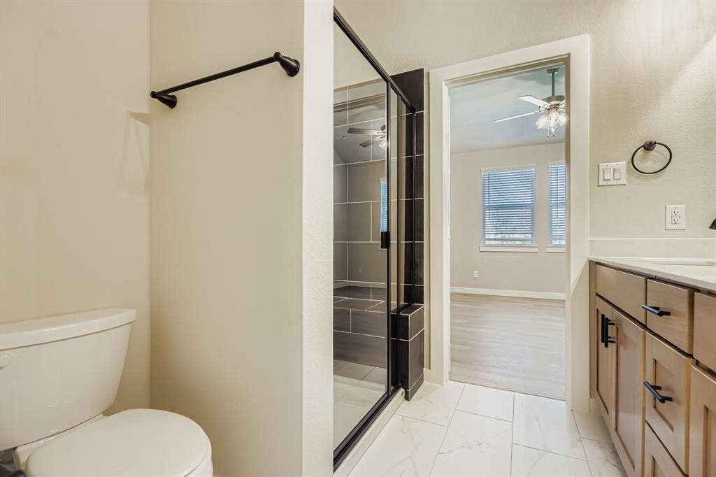 1204 Pebblebrook Drive, McKinney, TX 75071 Bathroom featuring a glass-enclosed shower with large-format tile surround, light wood-tone cabinetry, matte black hardware, and stone-finish floor tiling