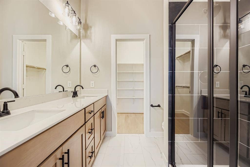 1204 Pebblebrook Drive, McKinney, TX 75071 Dual-sink vanity with light wood-finish cabinetry, white solid-surface countertops, and matte black fixtures, paired with a glass-enclosed shower featuring large-format tile surround and a walk-in storage area with built-in shelving