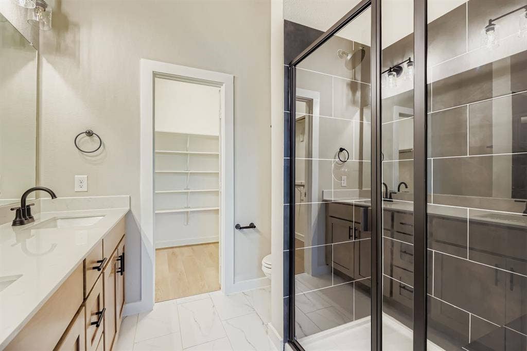 1204 Pebblebrook Drive, McKinney, TX 75071 Dual-sink vanity with light-toned cabinetry and quartz countertops, walk-in shower with dark-toned tile surround and matte black hardware, and built-in shelving