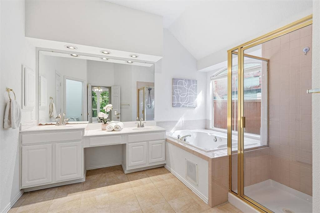 2902 Glen Hollow Court, McKinney, TX 75072 Dual-sink vanity with white cabinetry and expansive wall-to-wall mirror, built-in soaking tub, separate tiled shower enclosure with gold-tone frame, and arched window