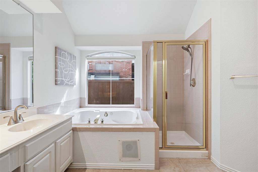 2902 Glen Hollow Court, McKinney, TX 75072 Bathroom featuring a drop-in soaking tub, a glass-enclosed shower with gold-tone framing, a double-basin vanity with a light-toned countertop, and arched window detailing