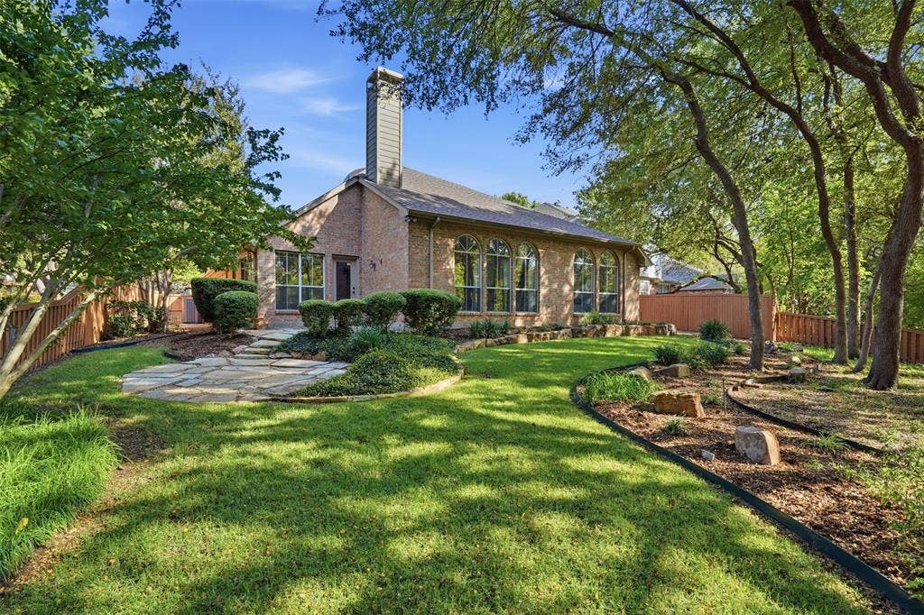 2902 Glen Hollow Court, McKinney, TX 75072 Brick exterior featuring arched windows, a stone-paved patio, mature trees, and a landscaped lawn with rock garden borders