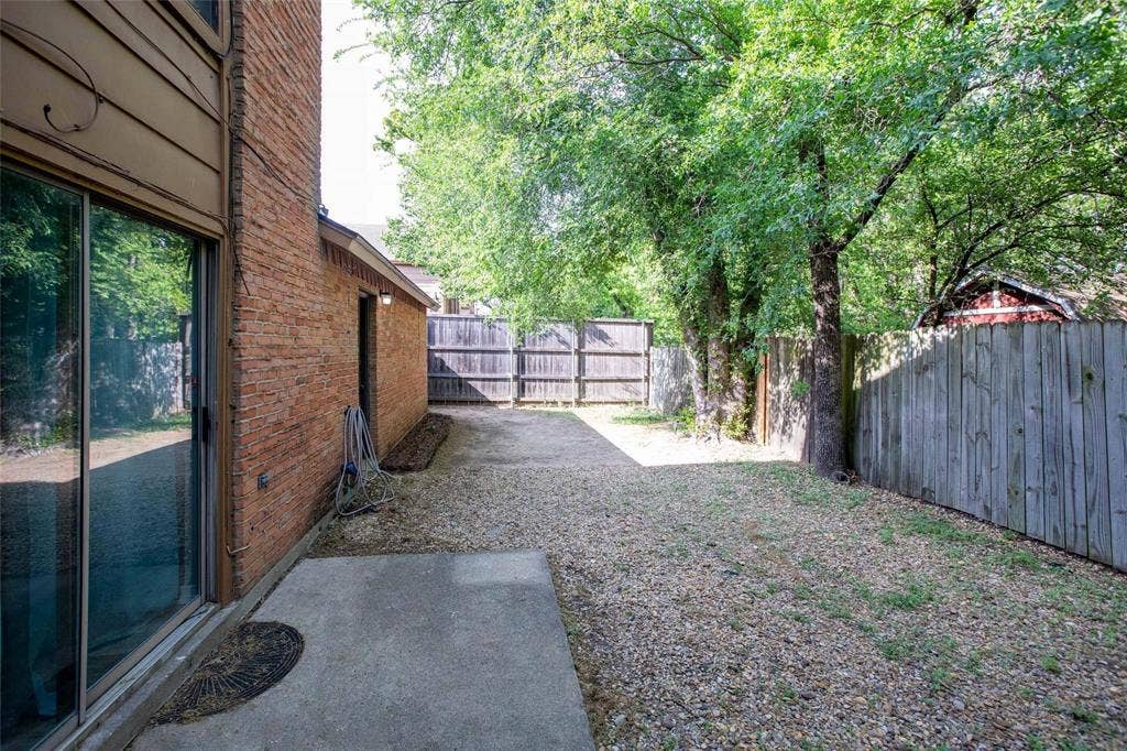 Private side yard featuring a brick exterior wall, concrete patio slab, gravel ground cover, wood privacy fencing, and mature canopy trees