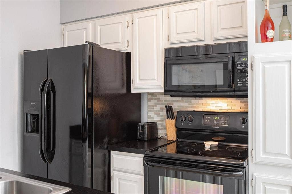 Kitchen featuring white shaker-style cabinetry, black appliances including a built-in microwave and range, dark countertops, and a glass-tile mosaic backsplash