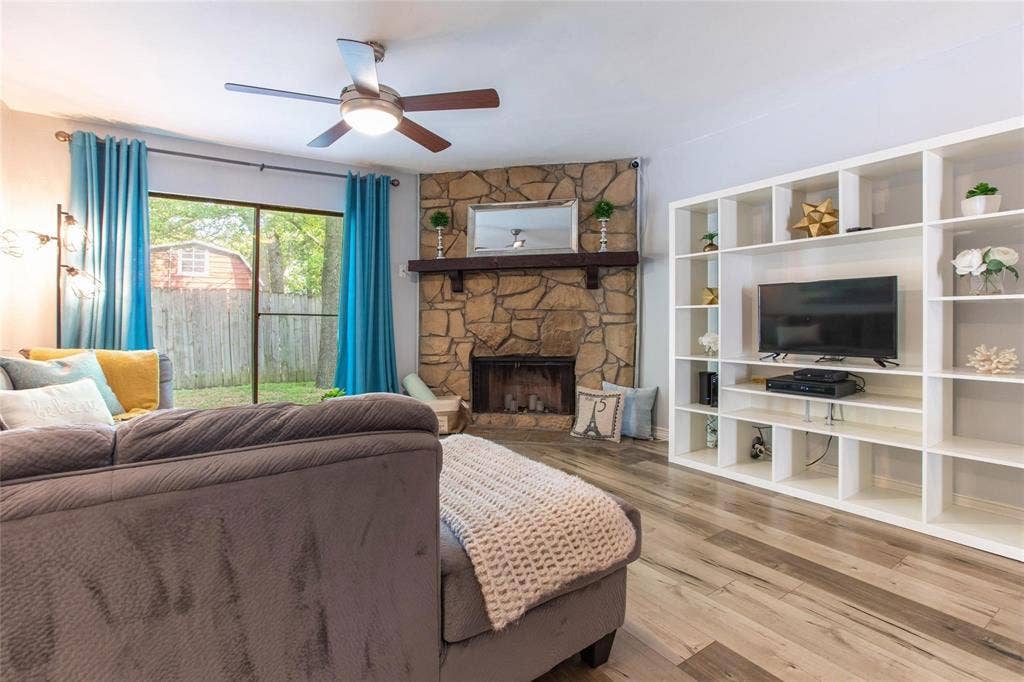 Floor-to-ceiling stone fireplace with a wooden mantel, wood-finish flooring, built-in white shelving unit, and a ceiling fan