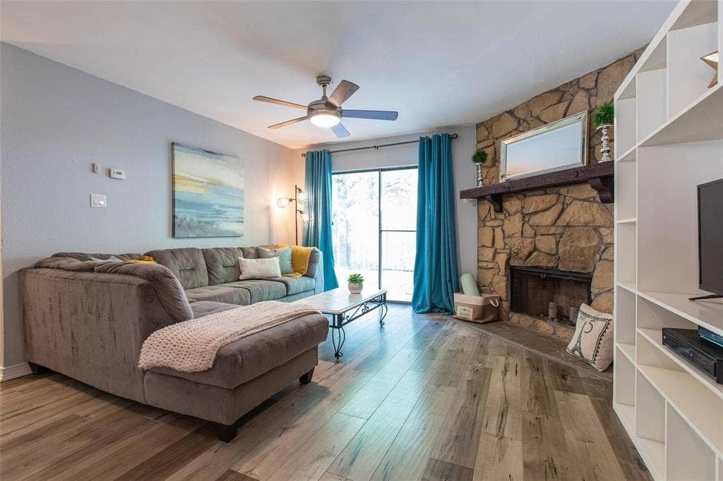 Living area featuring wood-finish flooring, a floor-to-ceiling stone fireplace with a thick timber mantel, a ceiling fan with integrated lighting, and sliding glass patio doors