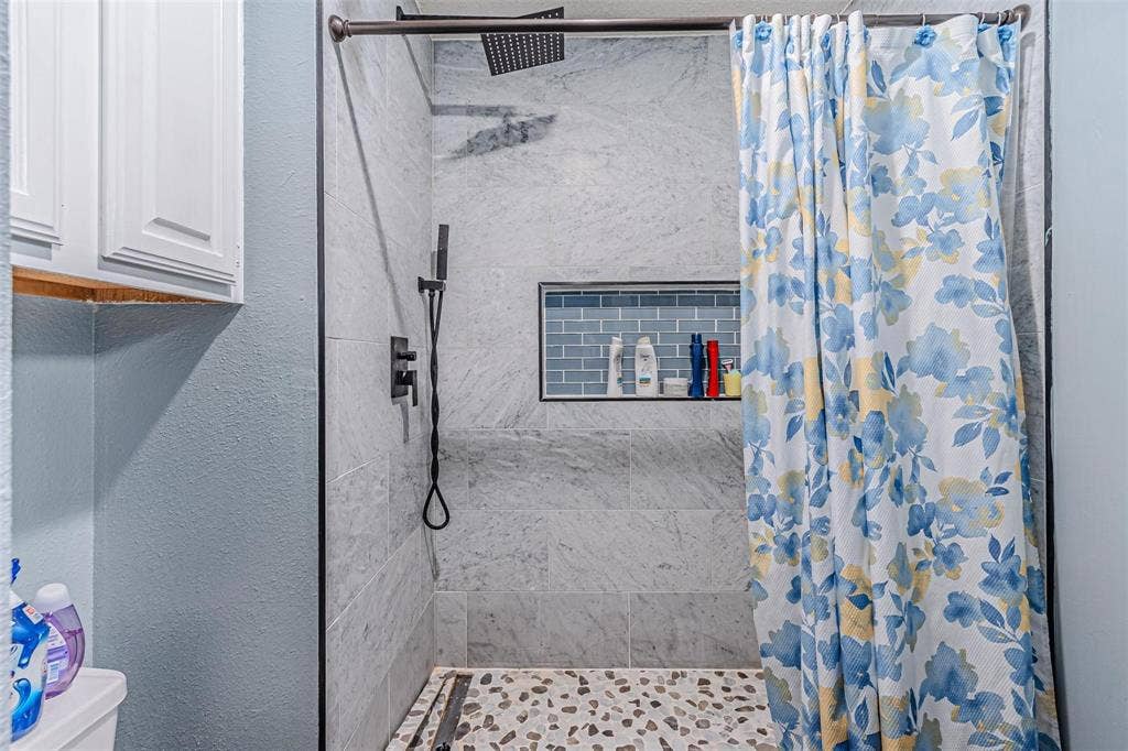 Walk-in shower featuring large-format stone-tile surround, blue subway-tile recessed niche, rainfall showerhead with matte black fixtures, and decorative pebble-stone flooring
