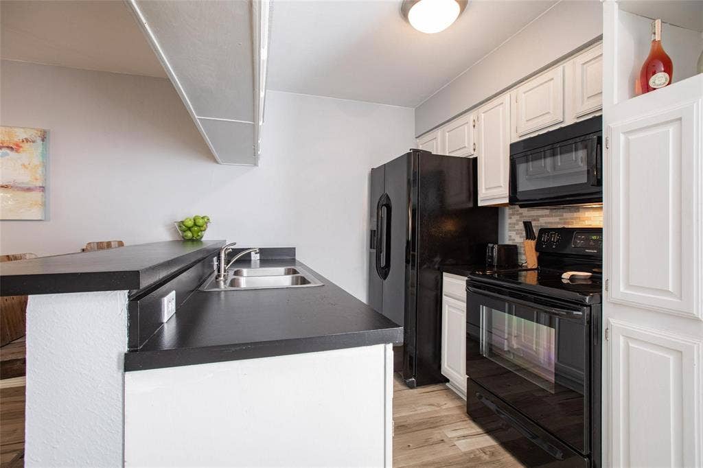 Kitchen featuring white shaker-style cabinetry, black appliances, dark solid-surface countertops, dual-basin stainless steel sink, and wood-finish flooring
