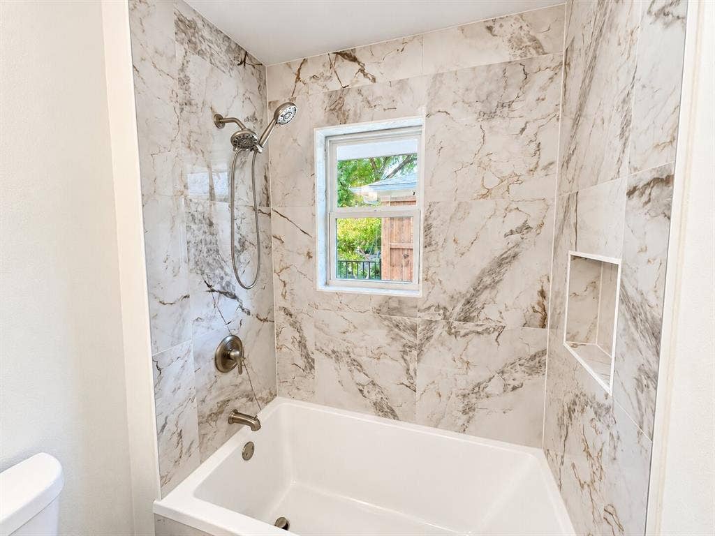 2551 Pinebluff Drive, Dallas, TX 75228 Bathtub surround featuring large-format stone-patterned tile, integrated recessed shelving, chrome handheld shower fixture, and a central window