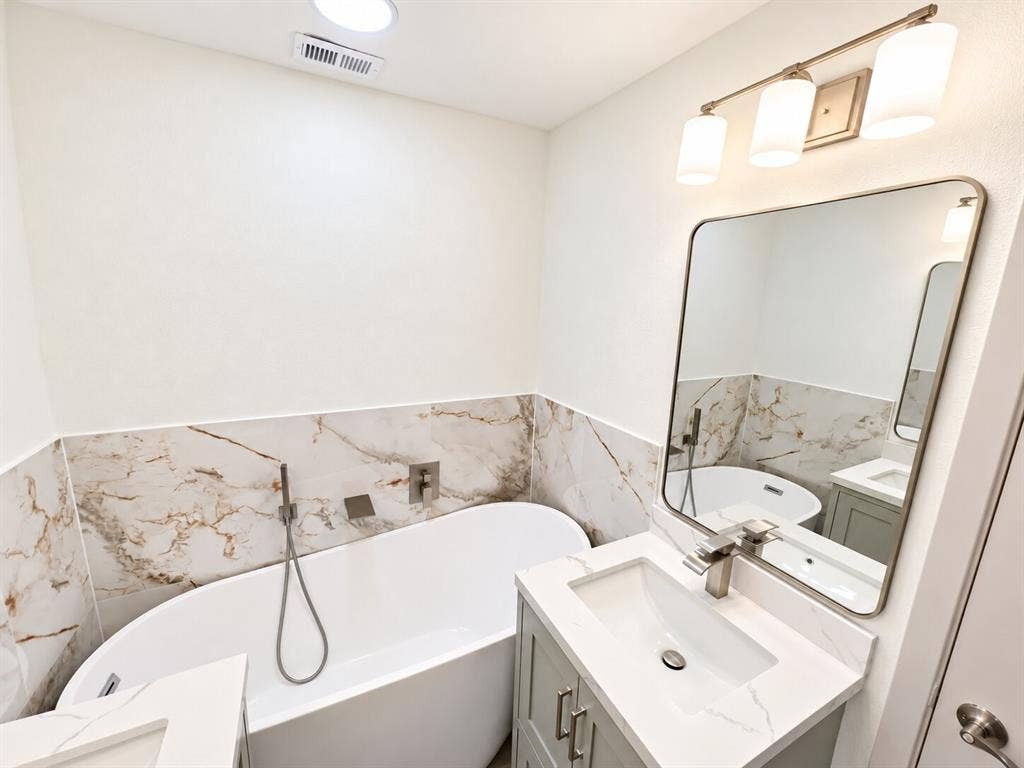2551 Pinebluff Drive, Dallas, TX 75228 Freestanding soaking tub featuring wall-mounted chrome fixtures, light-toned stone-finish wall tile, vanity with stone countertop and rectangular undermount sink, and minimalist three-light vanity fixture