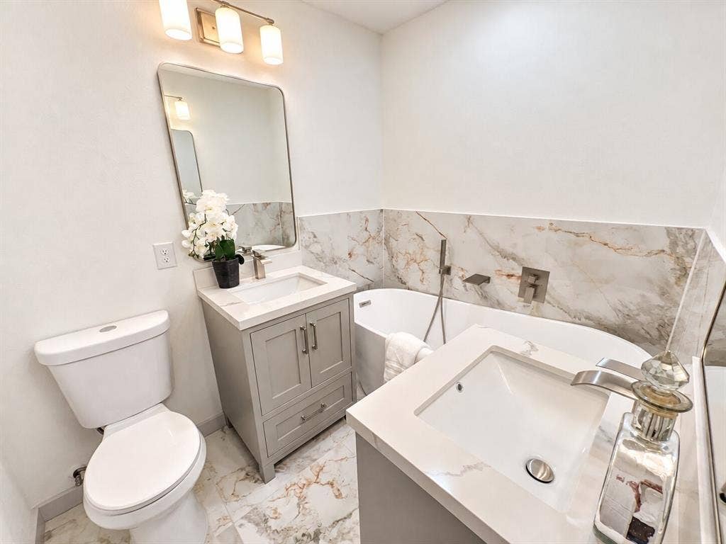 2551 Pinebluff Drive, Dallas, TX 75228 Bathroom featuring dual grey vanities with solid-surface countertops, soaking tub with wall-mounted hardware, floor-to-ceiling stone-patterned tile wall cladding, and oversized polished stone-patterned floor tiling
