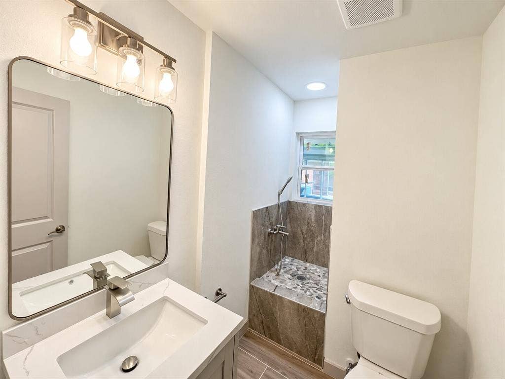 2551 Pinebluff Drive, Dallas, TX 75228 Bathroom featuring a vanity with a white countertop and integrated sink, a rectangular mirror with rounded edges, a three-light fixture with cylindrical glass shades, and a shower area finished with stone-textured wall tile and stone pebble flooring