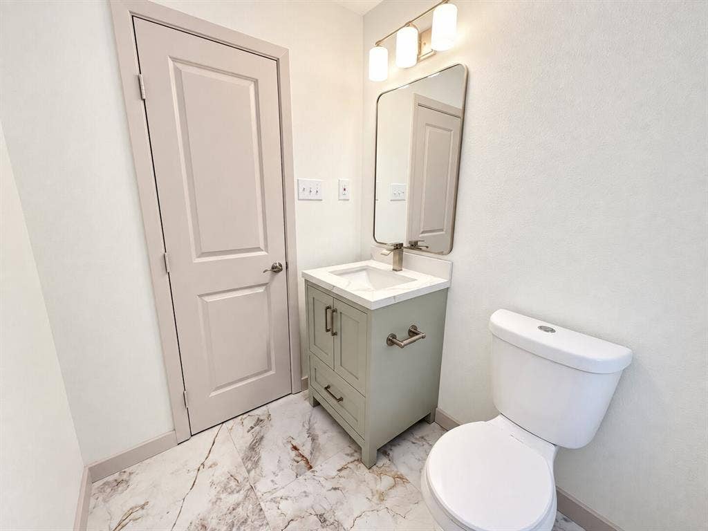 2551 Pinebluff Drive, Dallas, TX 75228 Single-sink vanity with a stone-surface countertop, rectangular undermount basin, rounded-edge wall mirror, three-bulb vanity light, and patterned tile flooring
