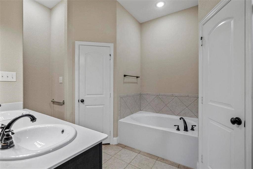 Deep-basin soaking tub with a tile surround, dual-sink vanity with a solid-surface countertop, recessed ceiling lighting, and ceramic tile flooring