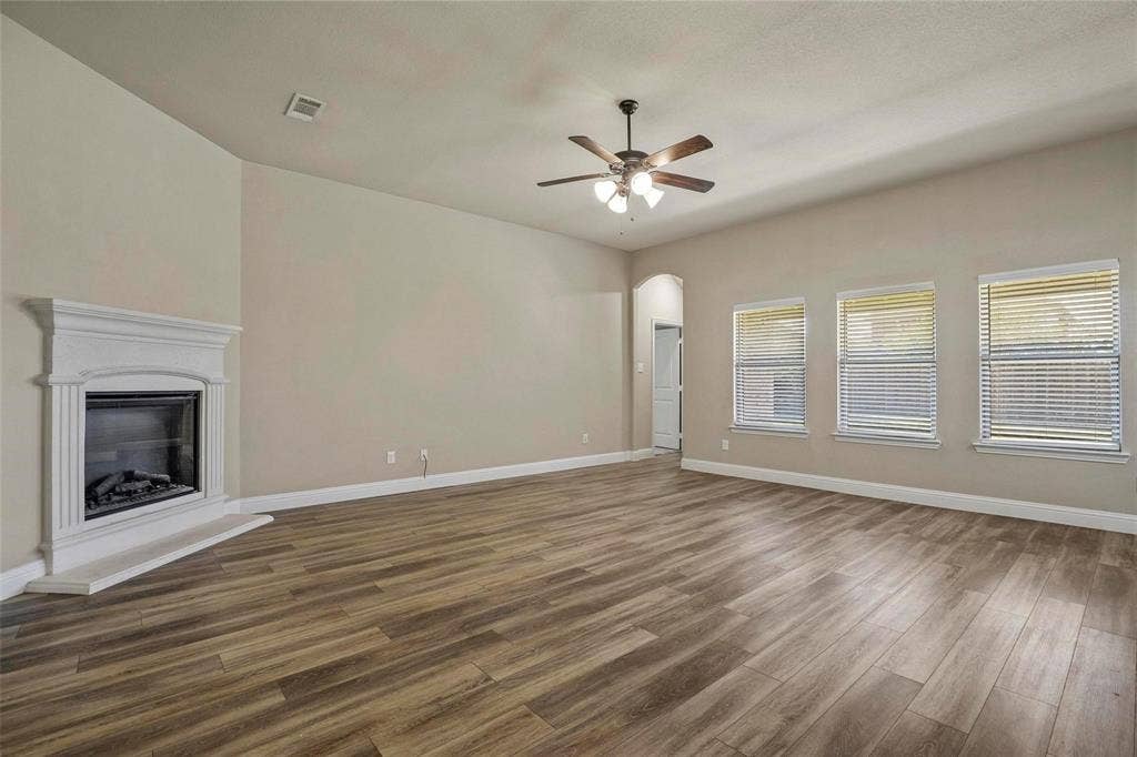 Open interior space featuring wood-finish flooring, a white mantel fireplace, three windows with blinds, and a ceiling fan with integrated lighting