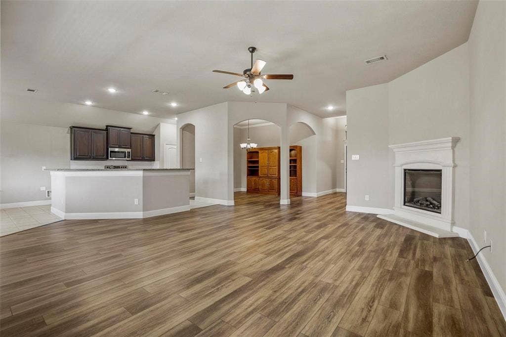 Open-concept interior featuring wood-finish flooring, a corner fireplace with a white mantel, arched architectural entryways, a kitchen with dark cabinetry and a raised breakfast bar, and recessed lighting