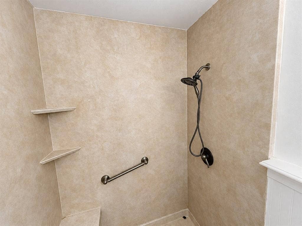 Shower stall featuring beige solid-surface surround panels, integrated corner shelving, a handheld shower fixture, and a horizontal metal grab bar