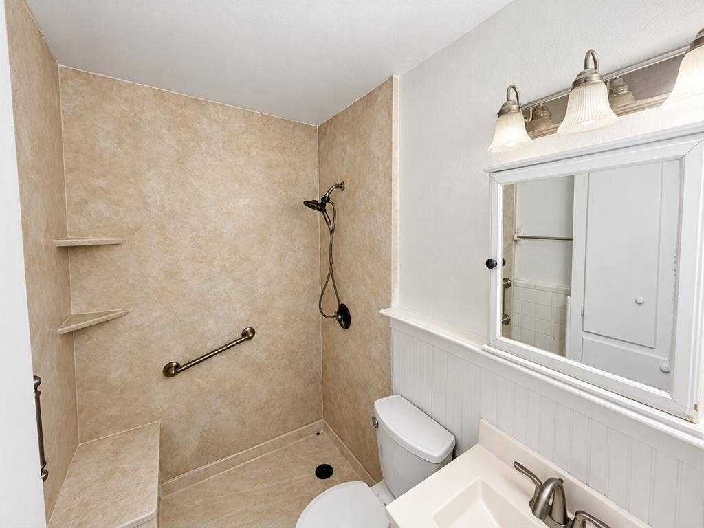Walk-in shower with integrated bench seating, solid-surface surround, and dual shower heads, complemented by a vanity with an integrated basin, beadboard wainscoting, and multi-bulb wall lighting
