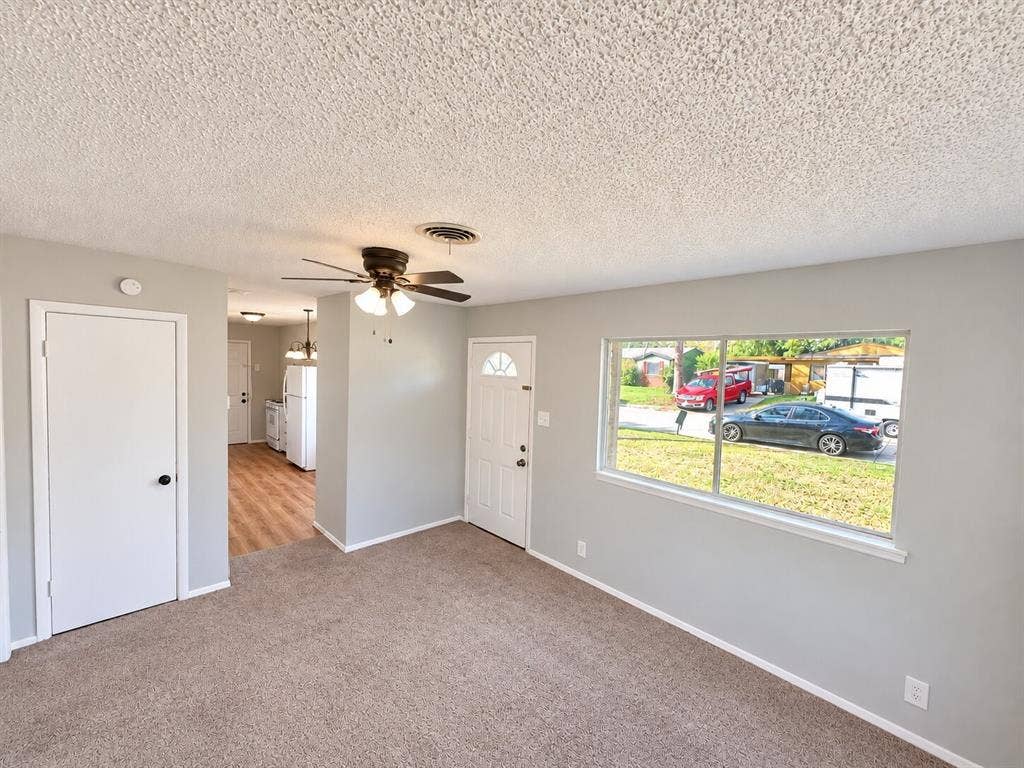 Living area featuring neutral wall paint, carpeting, a ceiling-mounted light fixture with fan, and a large window