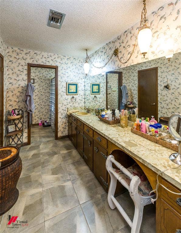 Extended vanity with stone-patterned countertop, integrated basin, wood cabinetry, tile flooring, and wall-to-wall mirror with pendant lighting