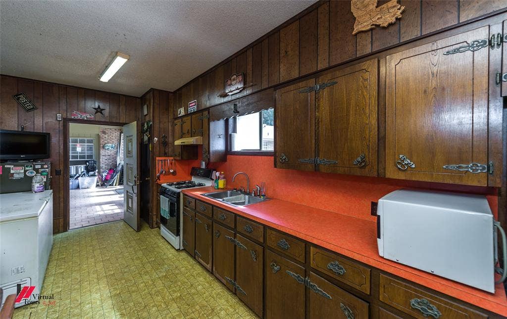 Kitchen featuring wood-panelled walls, ample wood cabinetry, solid-surface countertops, and a dual-basin stainless steel sink with a gooseneck faucet