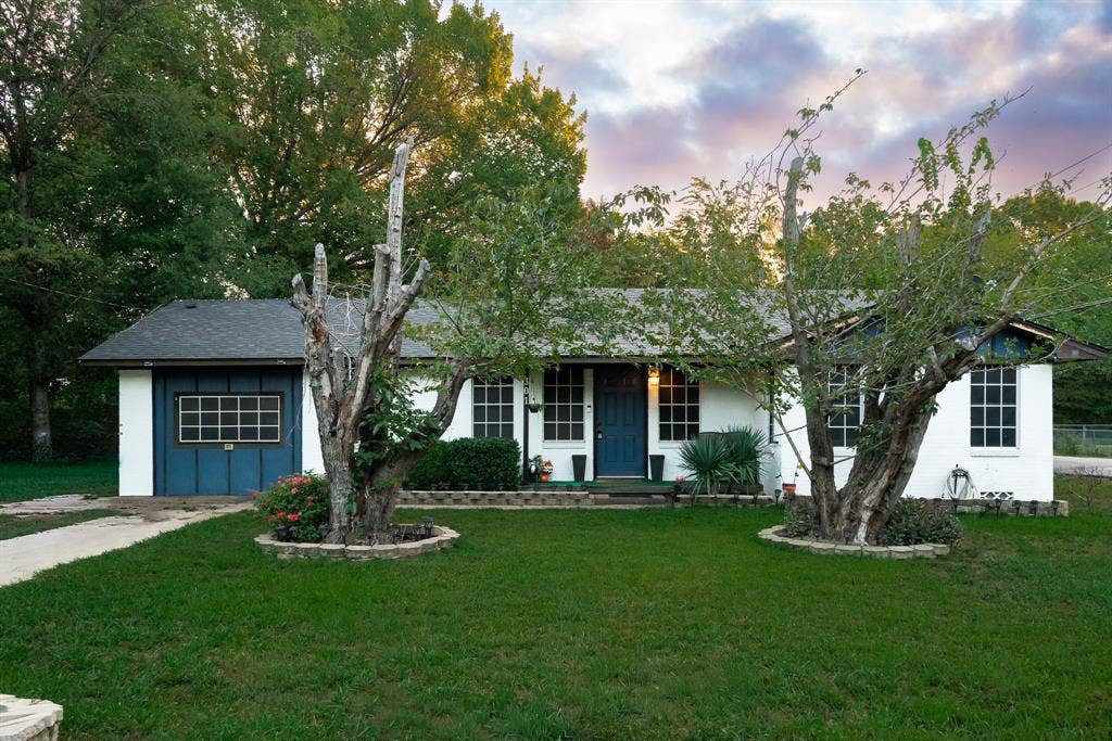 Single-story exterior featuring a white masonry façade, a blue single-car garage with an integrated window, a central blue front door, and manicured front lawn landscaping with stone-bordered garden beds