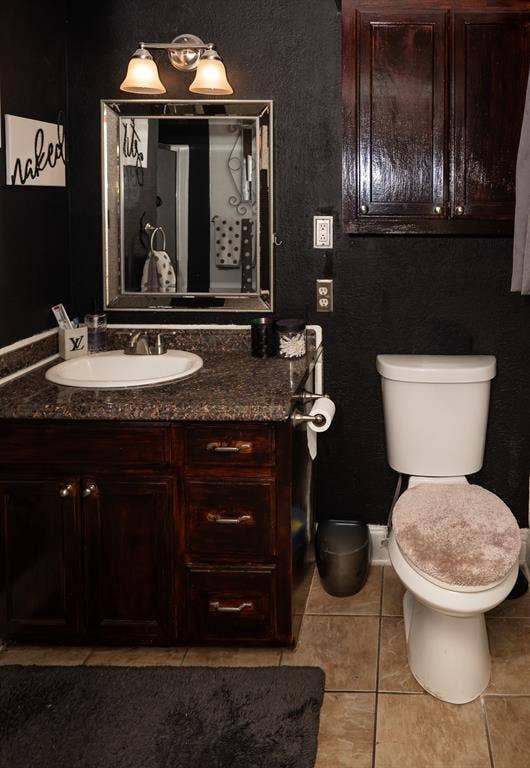Bathroom vanity featuring stained wood cabinetry, stone-finish countertop with an integrated basin, wall-mounted dual-light fixture, framed mirror, and upper wall-mounted storage cabinet