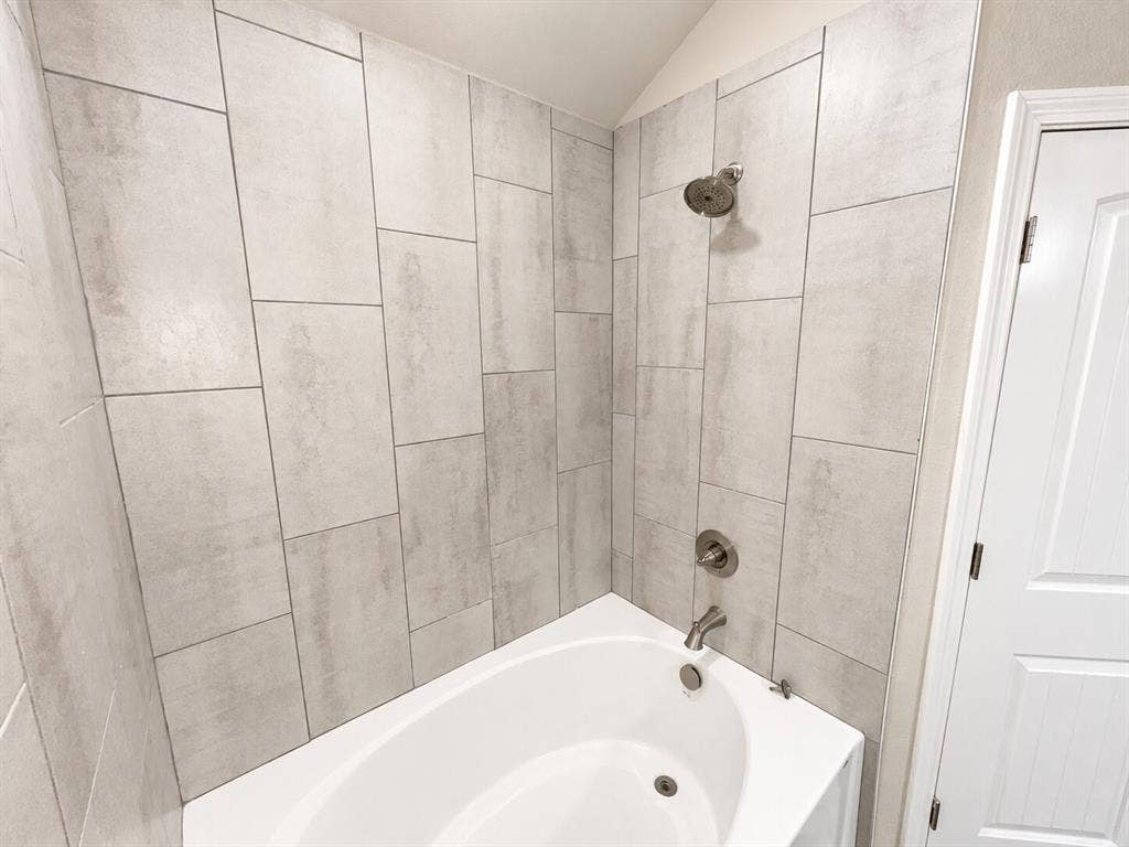 1306 Campbelton Drive, Temple, TX 76504 Bathtub featuring large-format light grey stone-patterned wall tile, brushed nickel shower head, matching tub faucet, and a multi-panel door