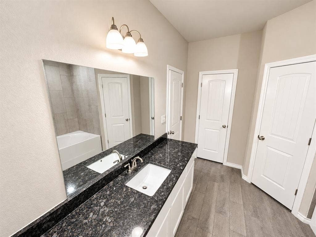 1306 Campbelton Drive, Temple, TX 76504 Dual-sink vanity with dark stone countertops, undermount rectangular sinks, wood-finish flooring, and a large wall-mounted mirror