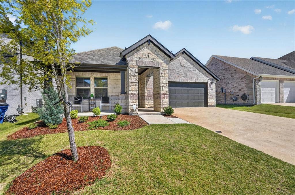 903 Edgehurst Drive, Princeton, TX 75071 Single-story residence featuring a stone and brick exterior, an attached garage with a dark panel door, a covered porch, a concrete driveway, and landscaped front yard with mulch beds