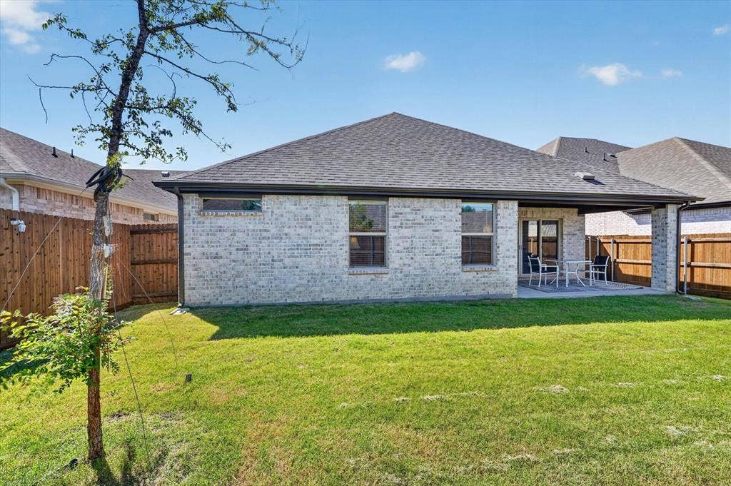 903 Edgehurst Drive, Princeton, TX 75071 Expansive green lawn with a covered patio, featuring a brick exterior with multiple windows, a dark shingle roof, and wood fencing