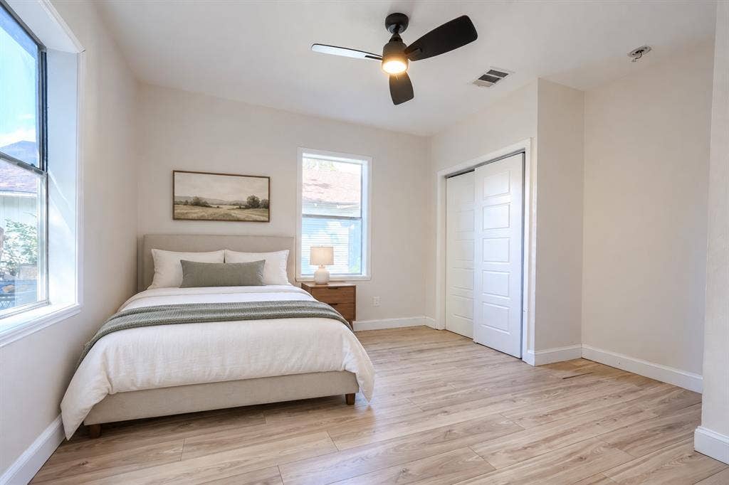 Neutral-toned interior featuring wood-finish flooring, dual windows, sliding closet doors with recessed paneling, and a matte black ceiling fan