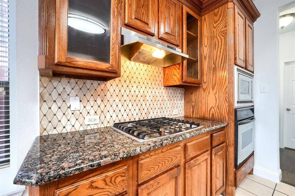 1127 Valley Vista Drive, Irving, TX 75063 Kitchen featuring custom wood cabinetry, natural stone countertops, a decorative diamond-patterned backsplash, a built-in stainless steel microwave and oven, and a gas cooktop