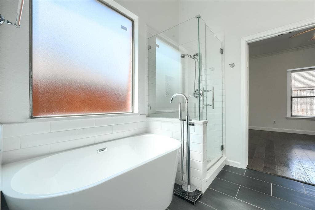 1127 Valley Vista Drive, Irving, TX 75063 Freestanding soaking tub with a floor-mounted chrome faucet, separate glass-enclosed shower featuring white subway tile, and dark large-format floor tile