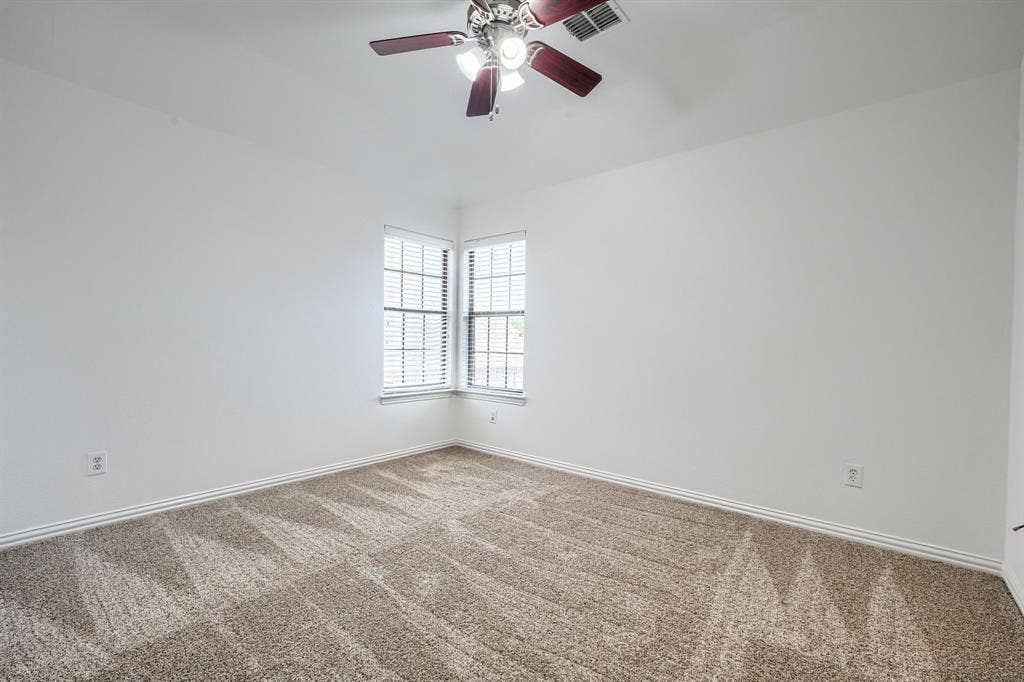 1127 Valley Vista Drive, Irving, TX 75063 Interior space featuring neutral wall tones, carpeted flooring, two corner-positioned windows with blinds, and a ceiling-mounted fan with integrated lighting