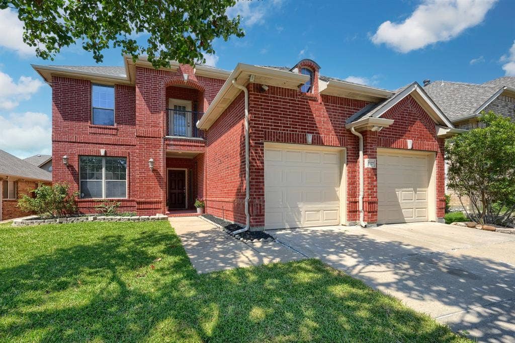 1127 Valley Vista Drive, Irving, TX 75063 Two-story red brick exterior featuring an arched entryway, a second-level balcony with metal railing, and an attached two-car garage