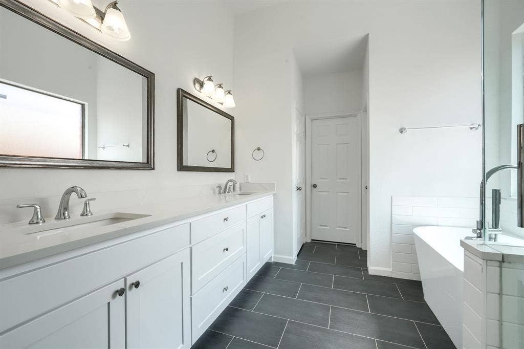 1127 Valley Vista Drive, Irving, TX 75063 Dual-vanity cabinetry with white finish, light-toned stone countertops, undermount sinks, framed mirrors, and large-format dark tile flooring