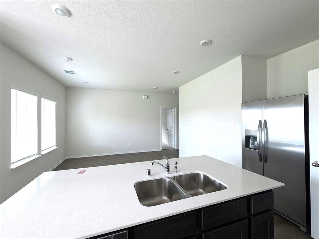 4017 Bronco Drive, Princeton, TX 75407 Kitchen island featuring a dual-basin stainless steel sink, polished chrome faucet, and solid-surface countertop, complemented by dark-toned cabinetry and a stainless steel refrigerator