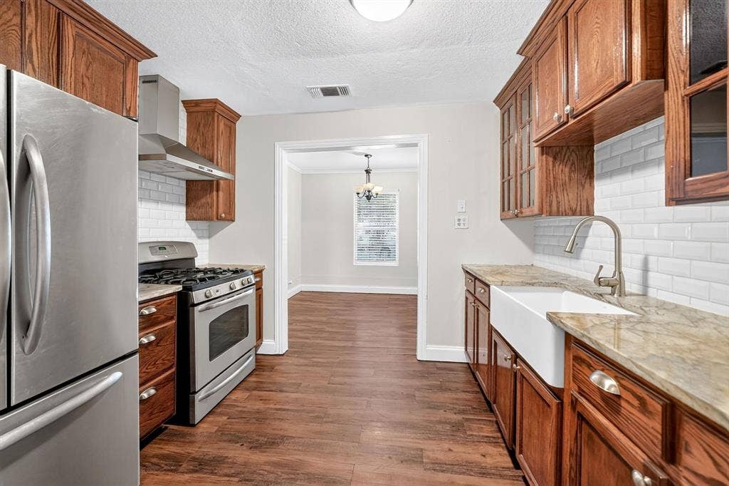 Kitchen featuring wood cabinetry, granite countertops, white farmhouse sink, white subway tile backsplash, and stainless steel appliances