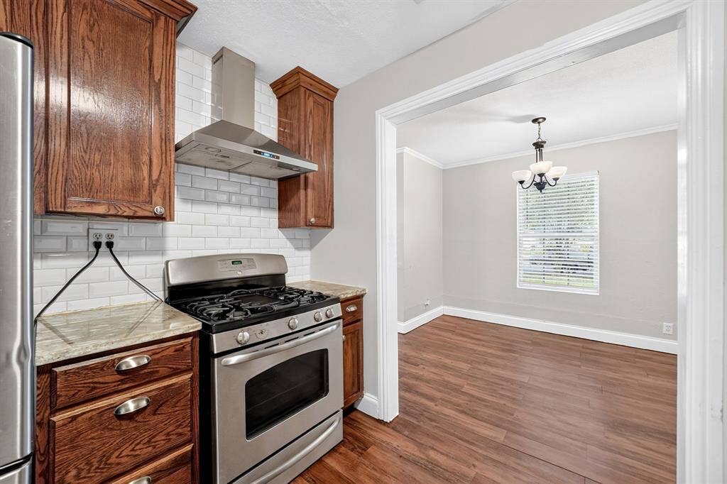 Kitchen featuring stained wood cabinetry, granite countertops, white subway tile backsplash, stainless steel gas range with overhead vent hood, and wood-finish flooring