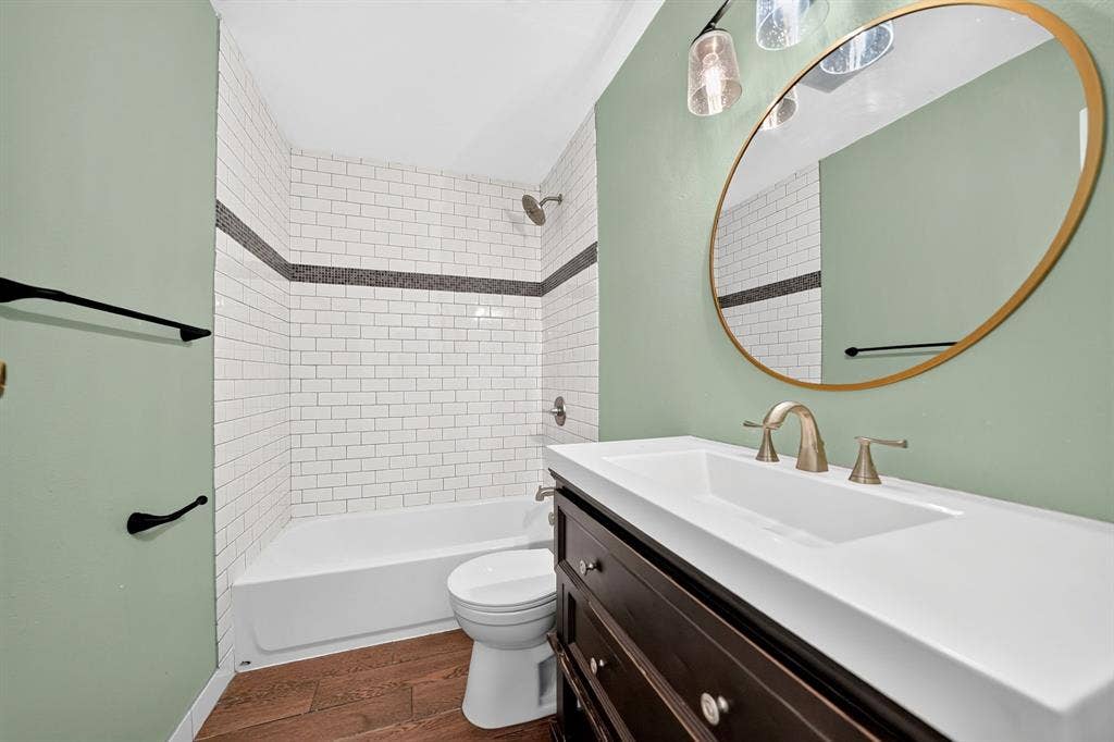 Bathroom featuring a dark wood-finish vanity with a white countertop, subway-tiled shower surround with a decorative mosaic accent stripe, wood-finish flooring, and a circular wall-mounted mirror