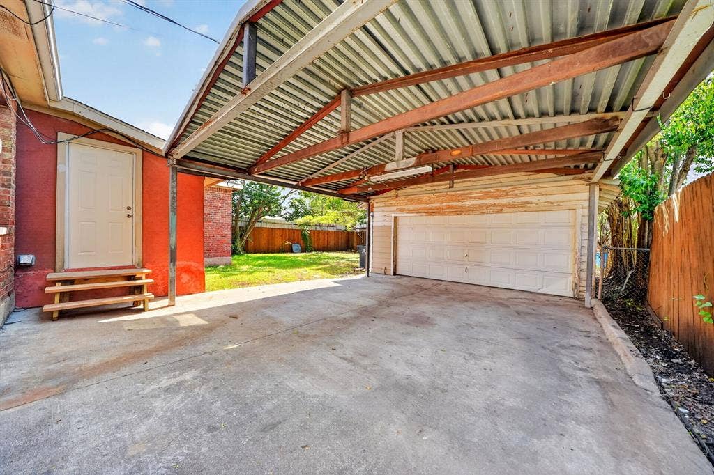 Concrete parking area with an attached metal-frame carport, featuring a white multi-panel garage door, exterior entry door with wooden steps, and a wooden privacy fence