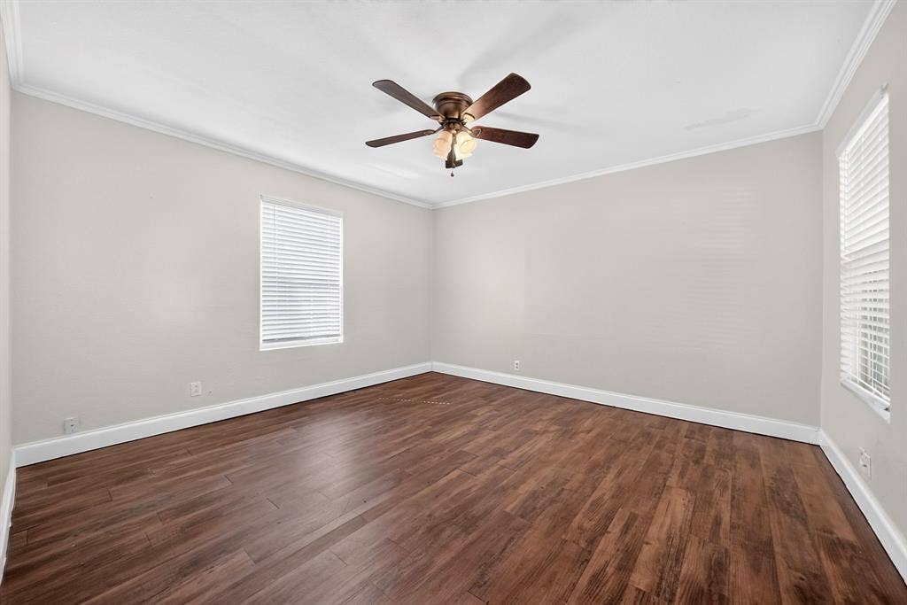 Neutral interior space featuring wood-finish flooring, crown molding, two windows with horizontal blinds, and a multi-blade ceiling fan with integrated lighting