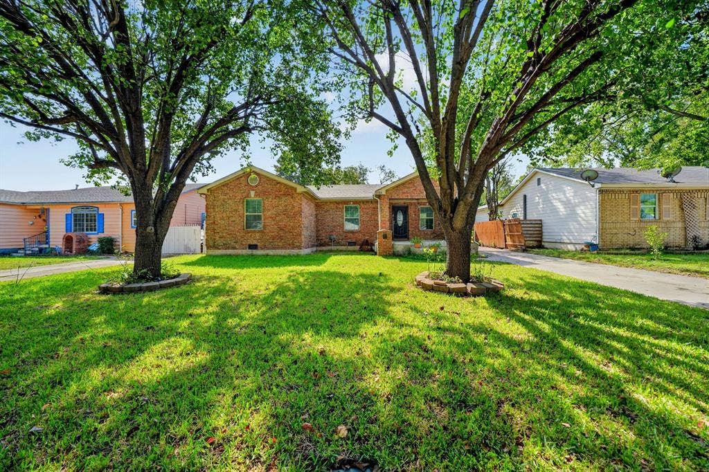 Brick exterior facade featuring a covered front entryway, concrete driveway, and established shade trees with stone-bordered landscaping