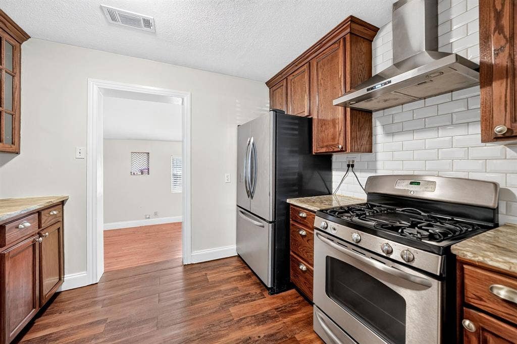 Kitchen featuring stained wood cabinetry, granite countertops, stainless steel appliances, a gas range with an overhead vent hood, and a white subway tile backsplash