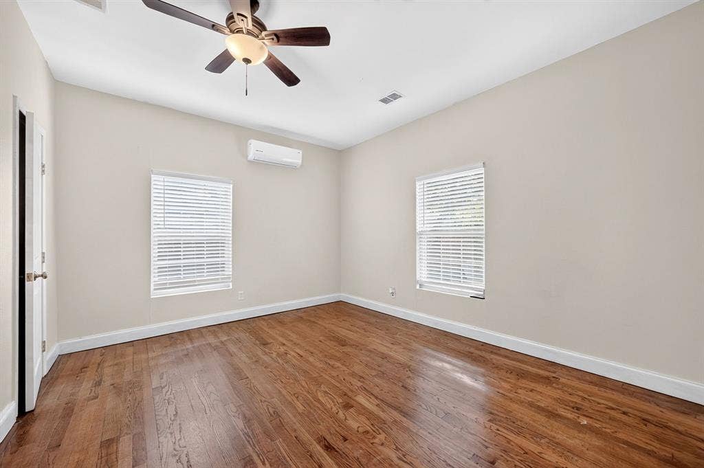 Room featuring wood-finish flooring, a wall-mounted climate control unit, dual windows with blinds, and an integrated ceiling fan with a light fixture
